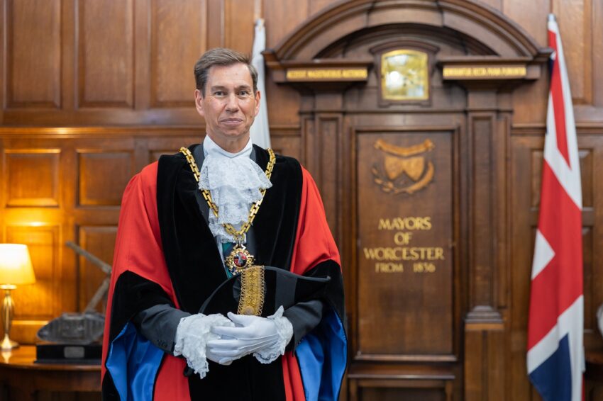 Mayor of Worcester Cllr Matt Lamb is red, black and blue Mayor Robes stood in front of a wood panelled wall with Mayor of Worcester text embossed on it in gold text.