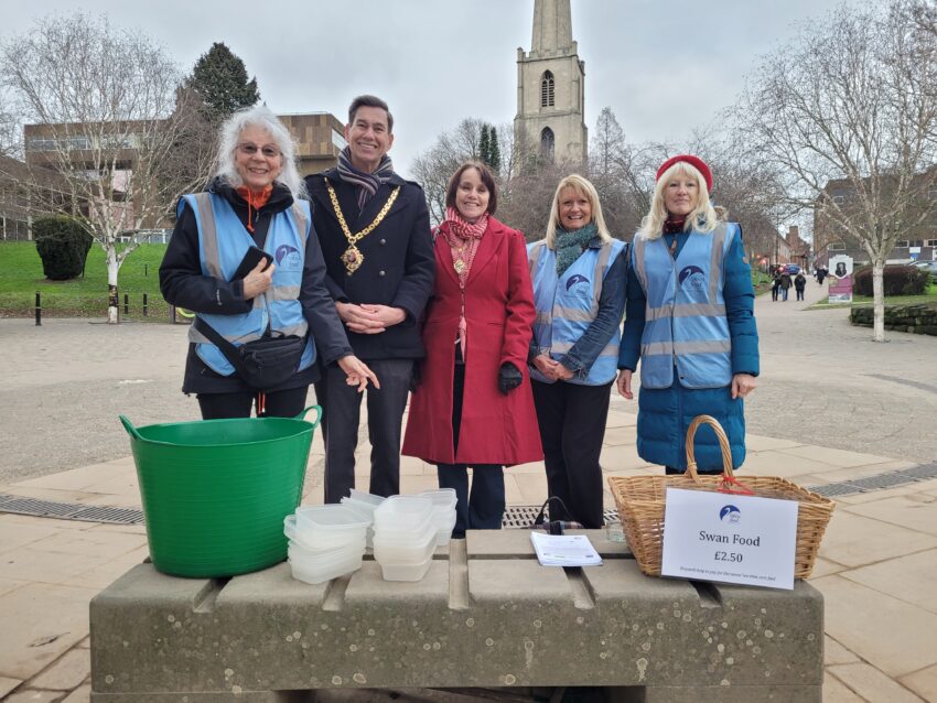 The Mayor and Mayoress standing with a group of women in front of a row of plastic containers and a basket with a sign attached that reads, "Swam food £2.50"