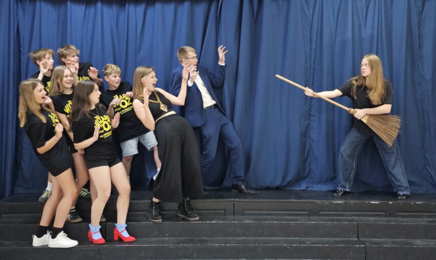 A theatre cast in rehearsals. A girl points a brromstick at a group of people, who cower in terror.